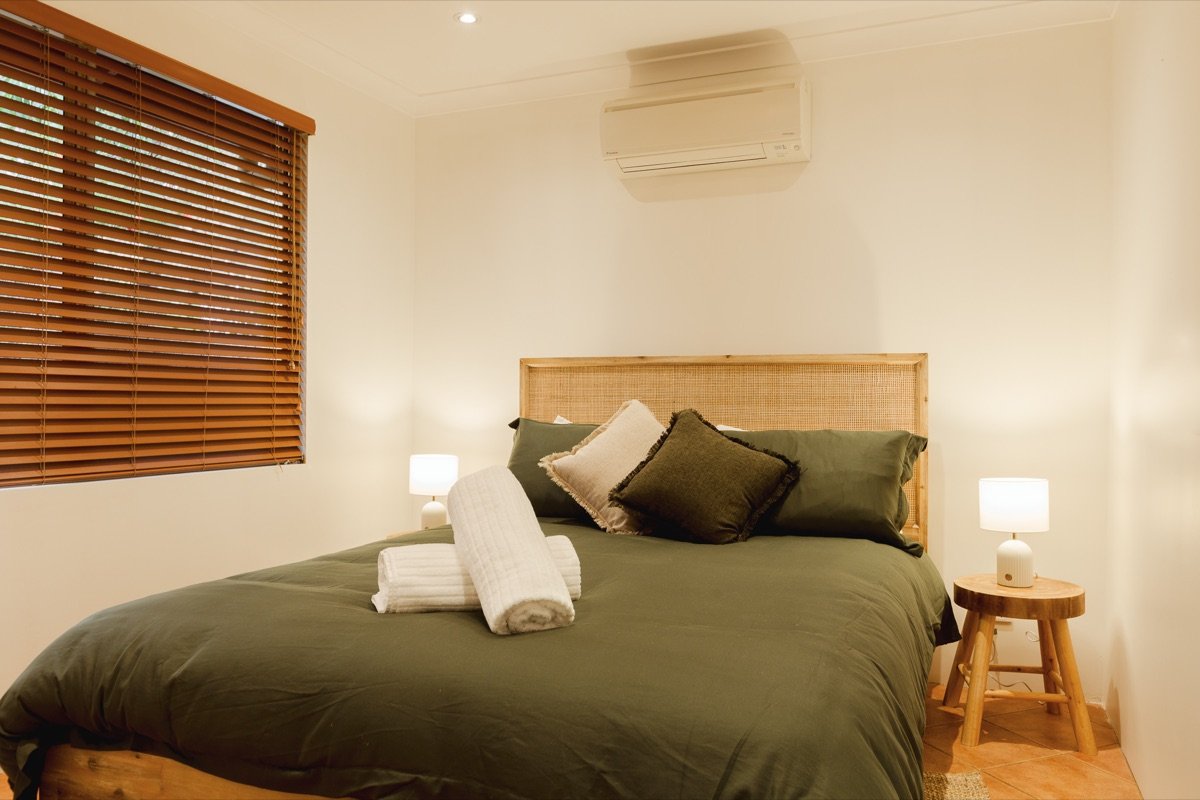 Second queen bedroom with deep green linen, rattan bedhead and timber blinds